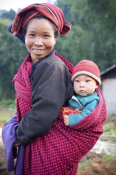 A mother beams with a warm smile, her baby snuggled securely against her back in a dotted red cloth. Her wrap is pulled high and tight, with the baby’s body held upright and close, legs tucked inside the fold of fabric. The baby peers out with wide,