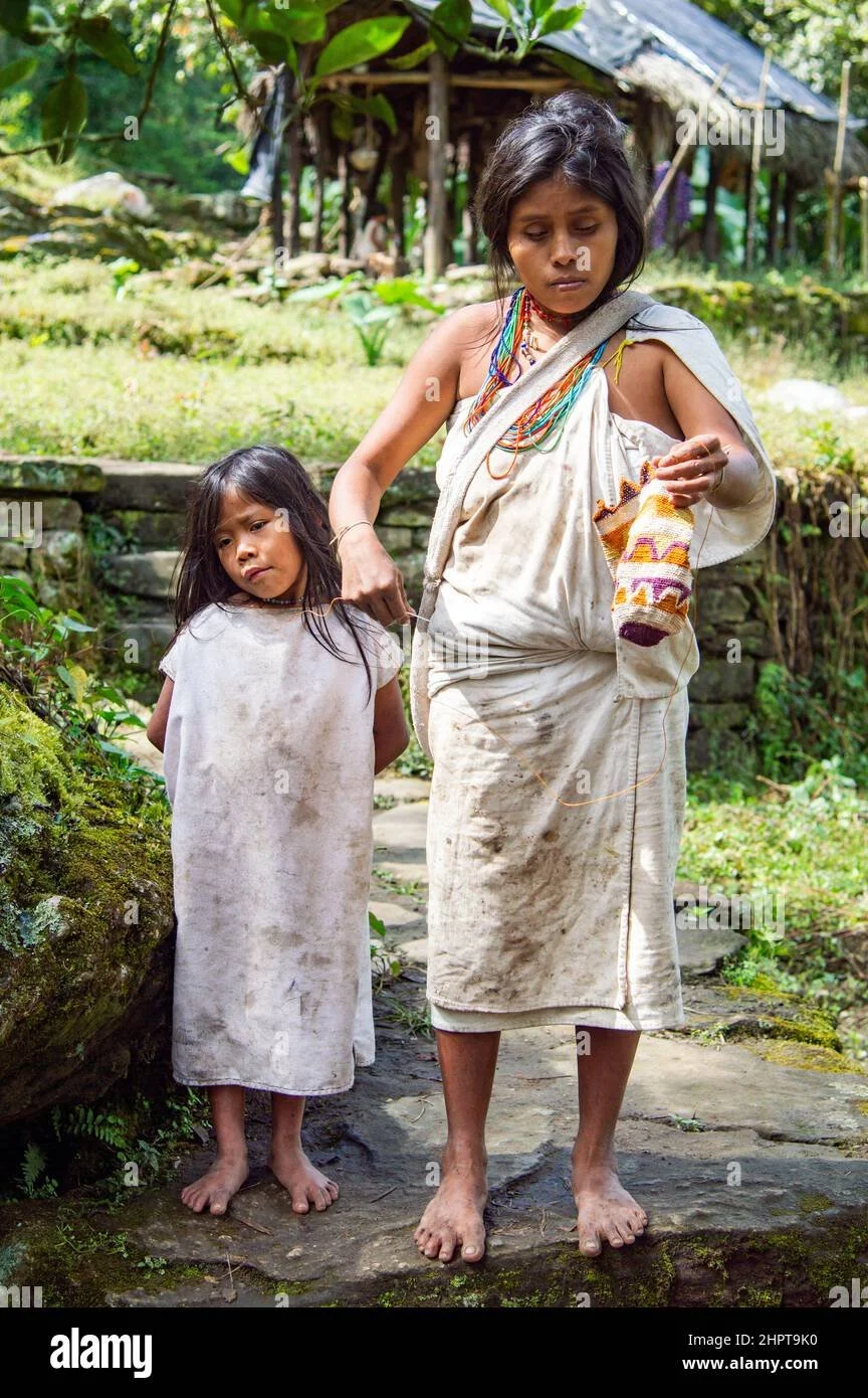 A Kogi mother and daughter stand barefoot along the moss-covered paths of Colombia’s Sierra Nevada, dressed in their distinctive handspun cotton garments. The woman holds a beaded piece of weaving-in-progress — perhaps a small mochila, the iconic bag