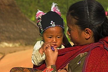 A mother gently tends to her baby, who is securely nestled in a deep red cloth sling wrapped across her front. The sling appears to support the child from beneath and around the torso, holding them upright and close against her body. The baby, dresse
