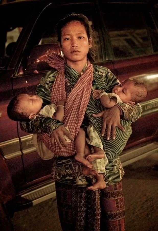 In this evocative night-time portrait, a mother stands holding her twin babies with a quiet, solemn strength. Each baby is nestled into a traditional krama scarf — the versatile checkered cotton cloth that is ubiquitous in Cambodia. One krama is loop