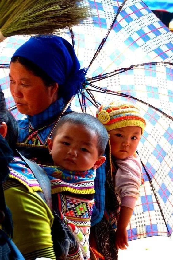 Two babies ride close on their mothers’ backs in the bustling colour and movement of a local market. Both are secured in traditional carriers that feature vibrant embroidery and structured panels, framed by broad shoulder straps and layers of woven c