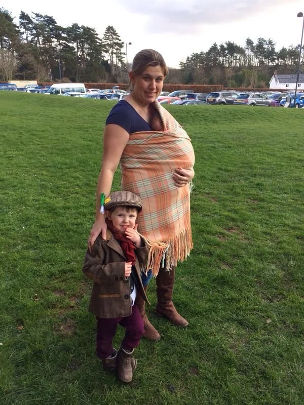 On a grassy field beneath a cloudy sky, a mother stands confidently with her two children — one at her side and one held close in a traditional woollen shawl. The wrap is worn in the iconic Welsh shawl carry, drawn diagonally across her body and knot