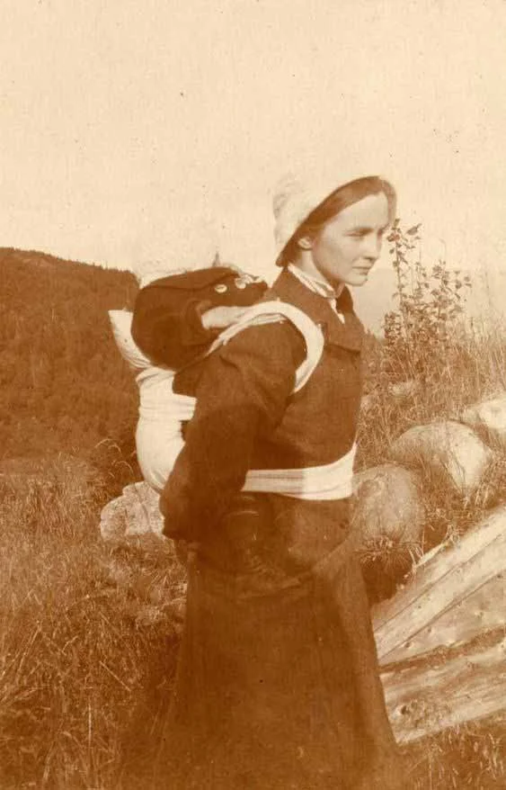 High on a grassy hillside, a Norwegian mother carries her child close in a soft back carrier, their bodies bound together with wide cloth straps that cross over her chest and waist. Her garments — a long dark dress, close-fitting bonnet, and crisp co
