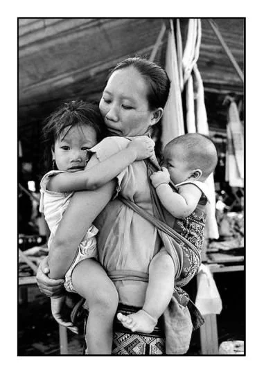 In this powerful black-and-white photograph, a mother carries two young children with quiet strength and tenderness. Her baby is tied securely to her back with a traditional cloth carrier — a wide, woven wrap that crosses over her chest and supports