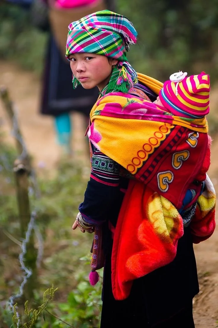Wrapped in colour and kinship, a young girl turns toward the camera, her expression calm but watchful. Her headscarf is a kaleidoscope of checks and tassels, wrapped high with the confident ease of tradition. Against her back, tucked deep into a brig
