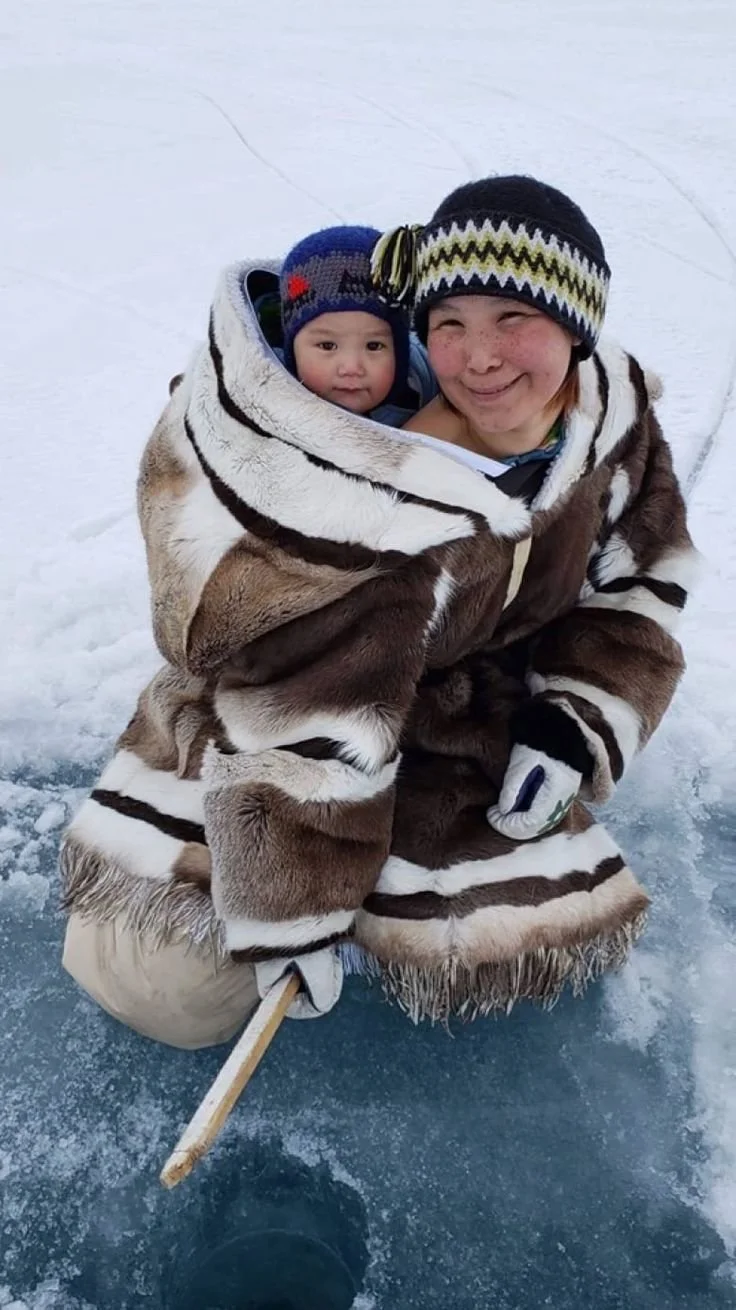 On the frozen surface of an icy lake, a mother kneels confidently with her baby snug against her back inside a traditional Inuit amauti. This amauti, crafted from beautifully patterned caribou or sealskin, surrounds them both in thick warmth, with th