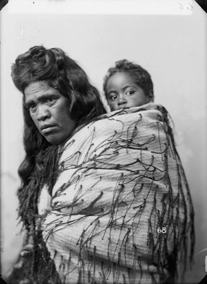A Māori woman turns slightly toward the camera, her gaze steady and strong, while a child peeks quietly over her shoulder from within the embrace of a traditional kākahu. The cloak is richly adorned with hukahuka — decorative flax fibre tassels that