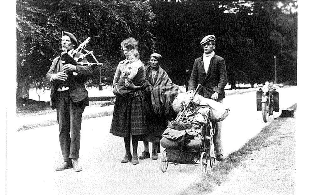 A family walks down a country road, their lives packed into a pram, their stories stitched into every step. At the front, a man plays the bagpipes, leading the way with rhythm and resolve. Beside him, a mother carries her child in arms. Another woman