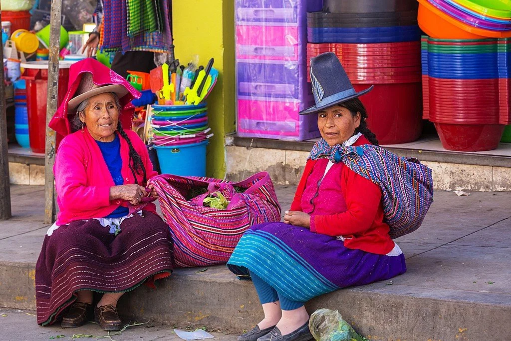 Two women rest on a stone step in a bustling Peruvian market street, their skirts and shawls a vivid blend of texture and tradition. The woman on the right wears her large carrying cloth — an aguayo — wrapped around her back in a familiar way, its we