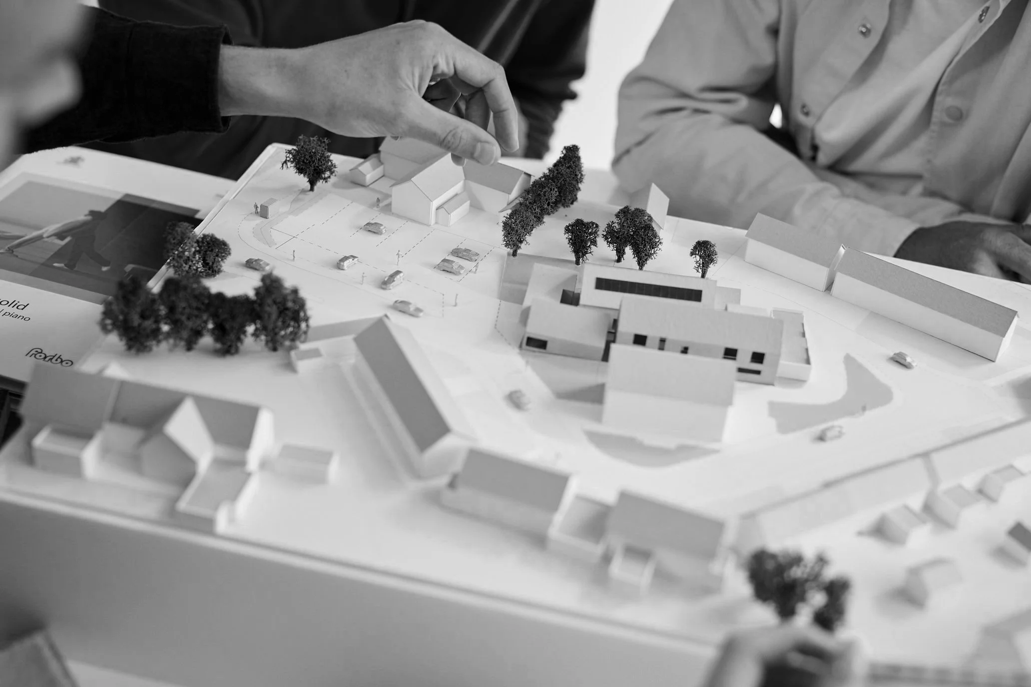 People examining a detailed architectural model of a residential development with trees and vehicles, in black and white.