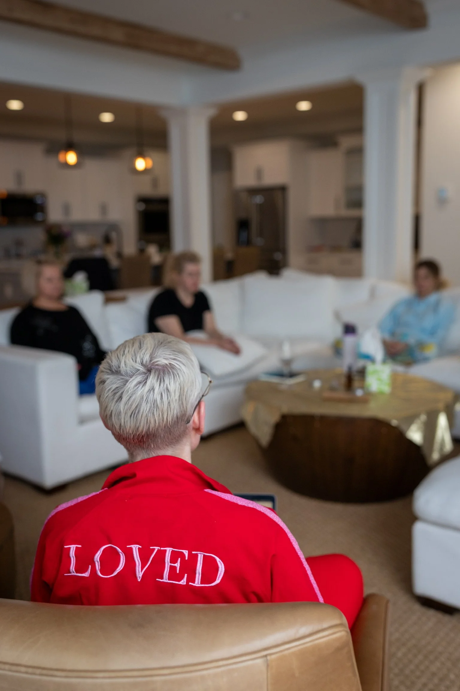 A person with short, light-colored hair wearing a red jacket with the word "LOVED" on the back, sitting on a beige chair in a living room, facing three women on a white sectional sofa, with a kitchen in the background.