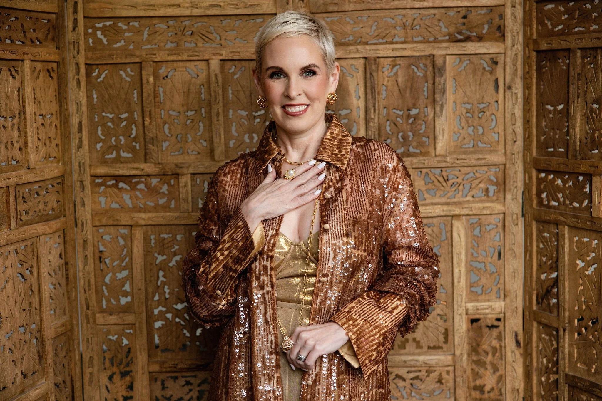 A woman with short blonde hair and blue eyes wearing a shiny, copper-colored, sequined jacket over a gold top, standing in front of a decorative wooden screen.