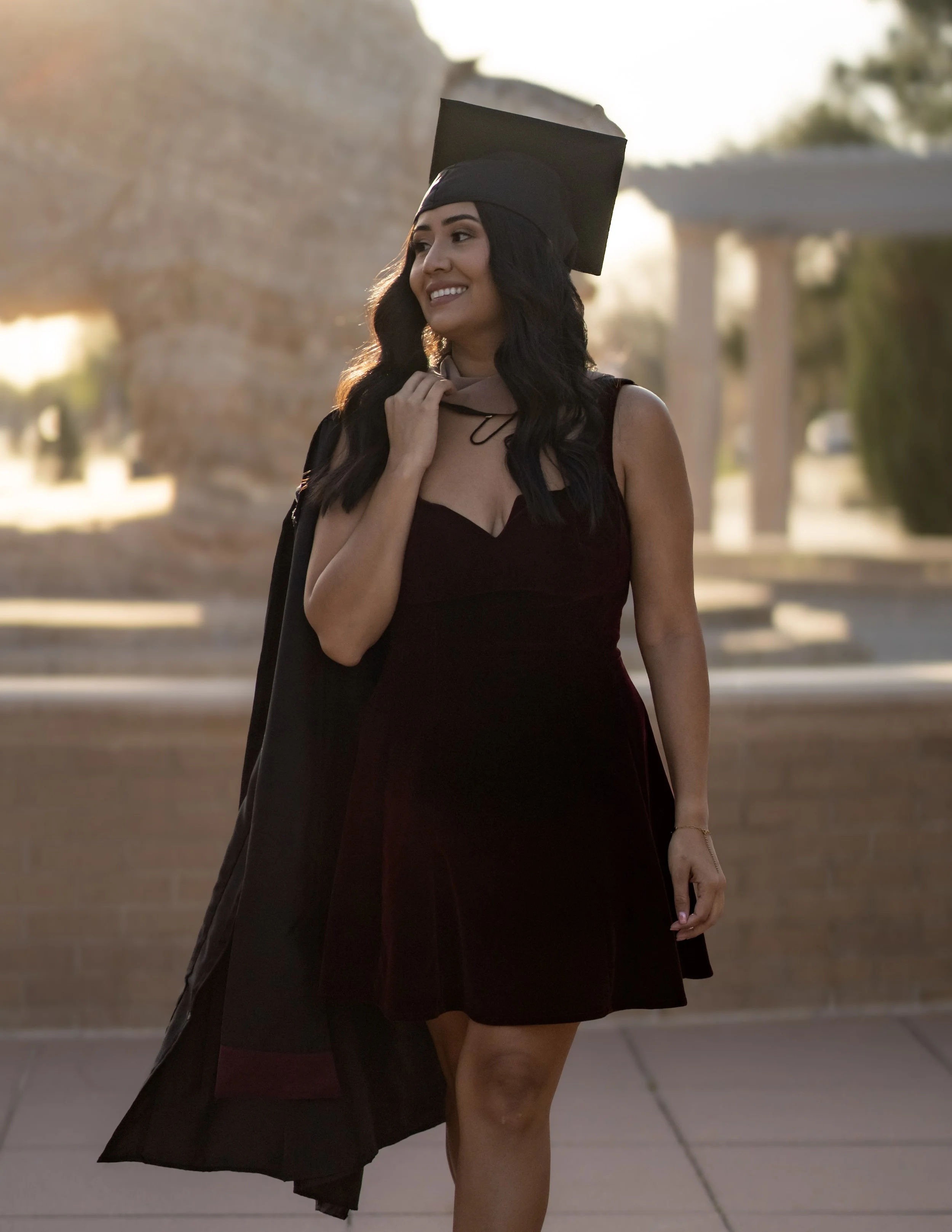 Young woman in a black graduation cap and dress standing outdoors during sunset, smiling, with a brick wall and trees in the background.