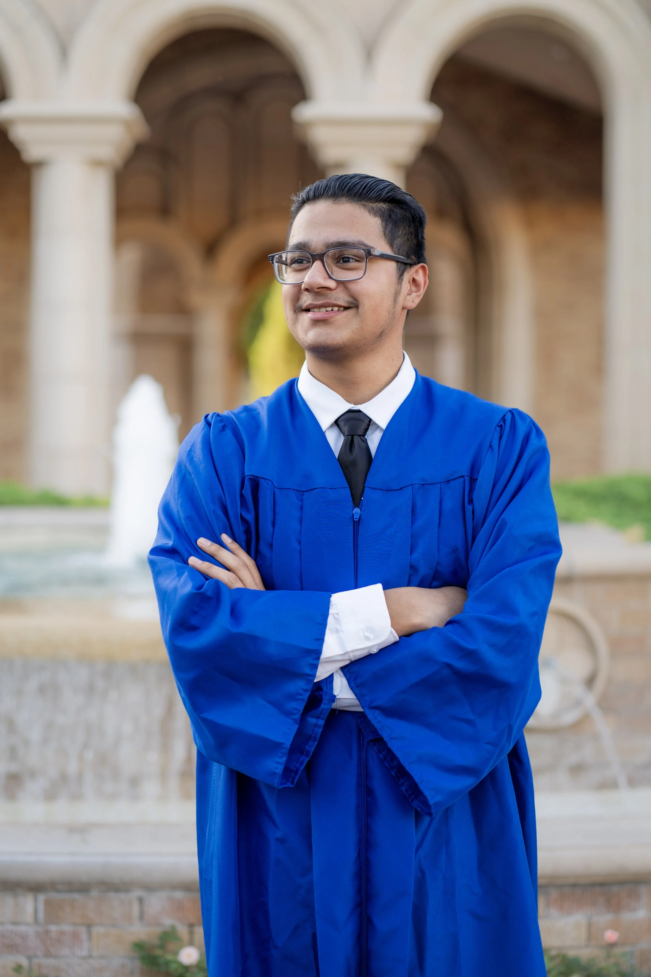 Young man in graduation gown and glasses standing outdoors with arms crossed, smiling, in front of arches and fountain.