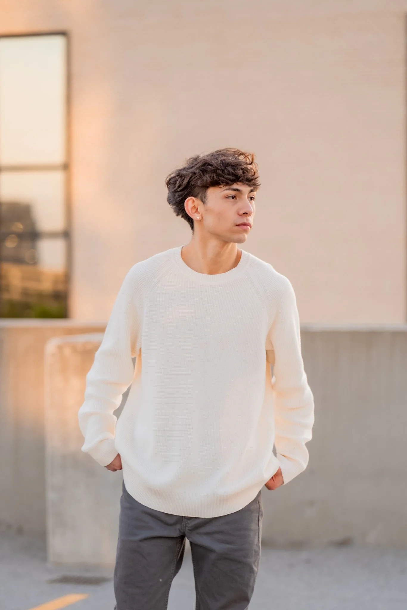 A young man with wavy dark hair, wearing a white sweater and gray pants, stands outdoors in front of a beige wall with a window, with one hand in his pocket, looking to the right.