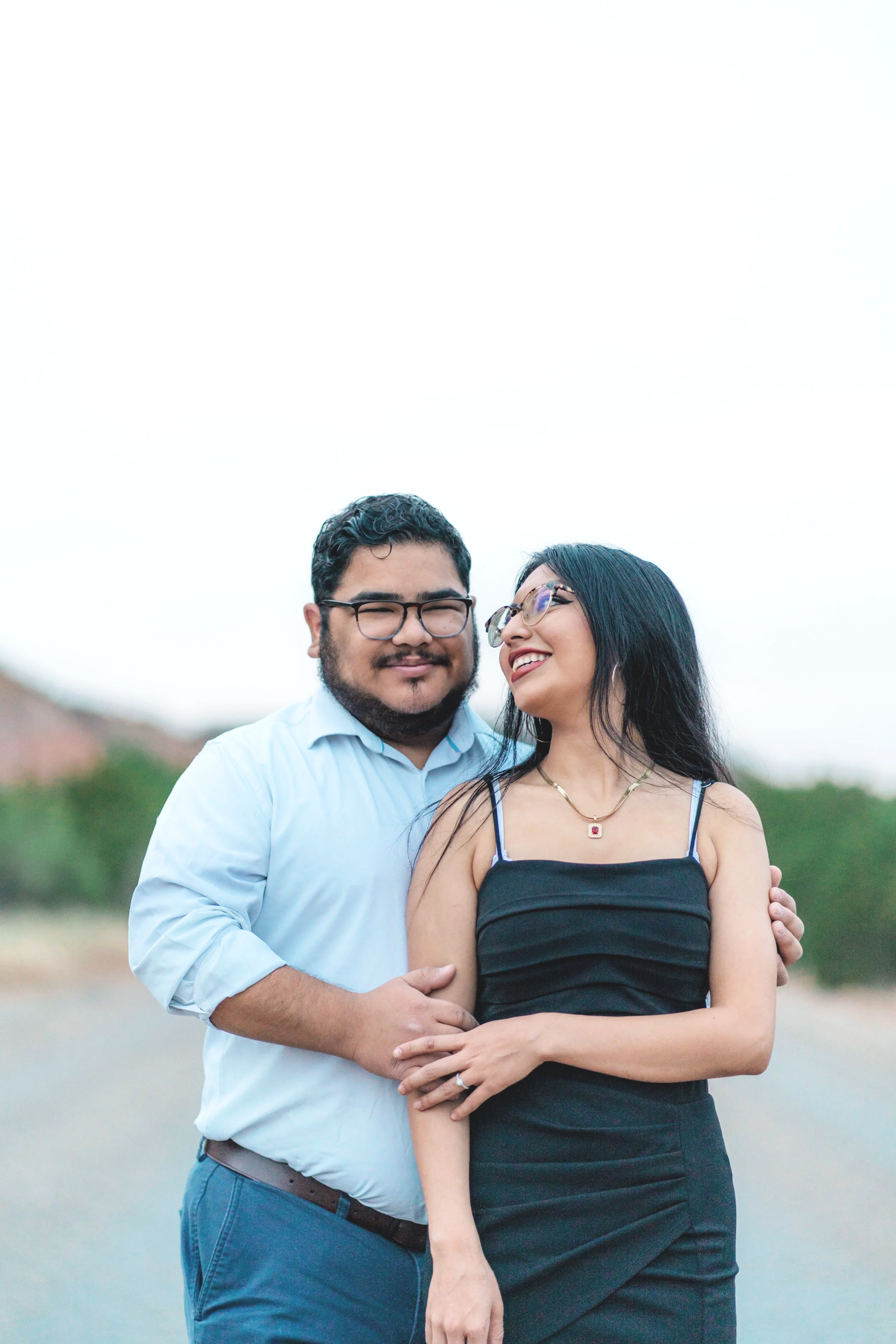 A happy couple stands closely together outdoors, with the man's arm around the woman's waist and both smiling. The man has glasses and a beard, wearing a light blue button-up shirt. The woman has long black hair, glasses, and is wearing a black tank 