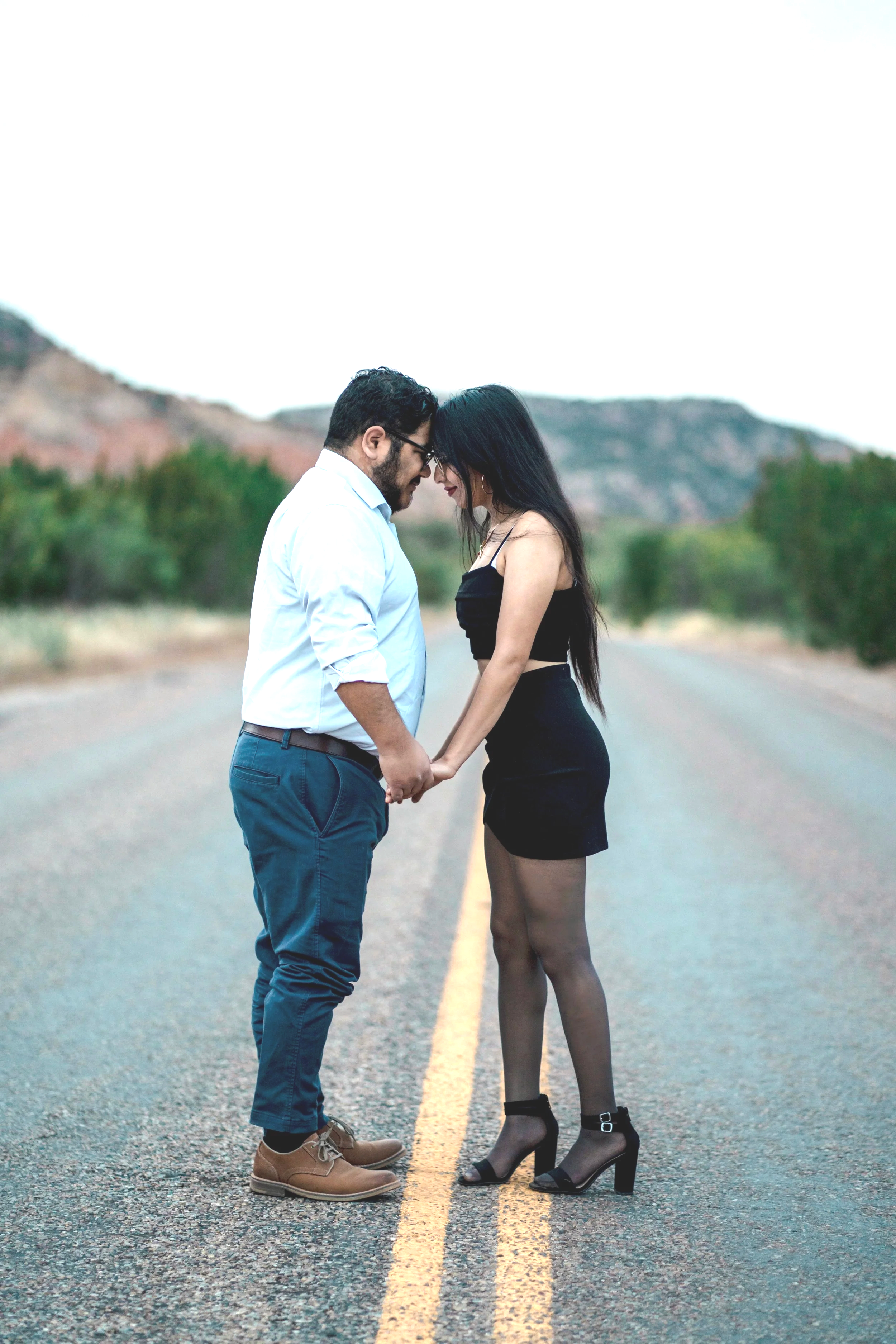 A man and woman stand close together on a deserted road with a mountain backdrop, touching foreheads and holding hands.