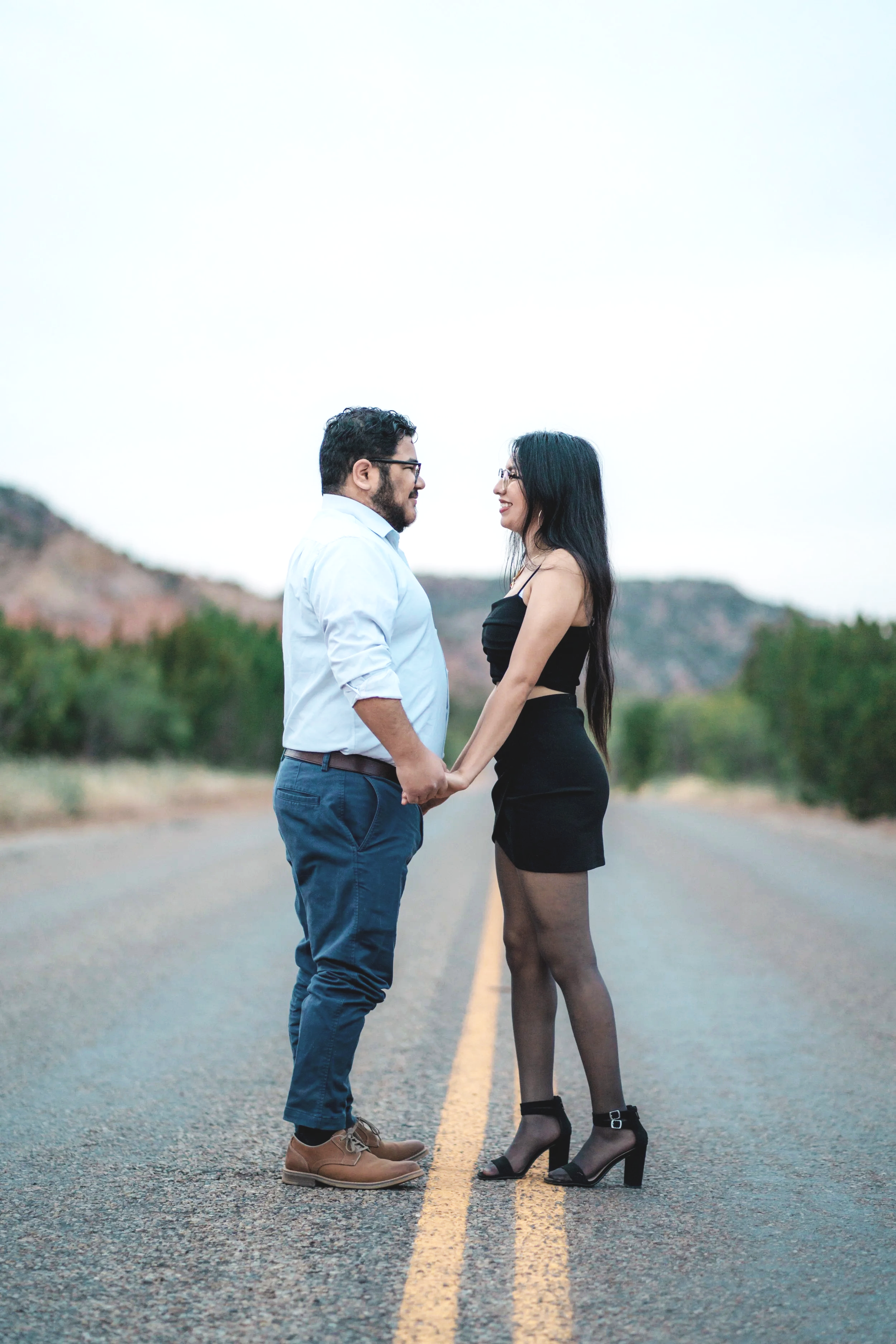 A man and woman standing face to face holding hands in the middle of a deserted road with mountains and trees in the background.