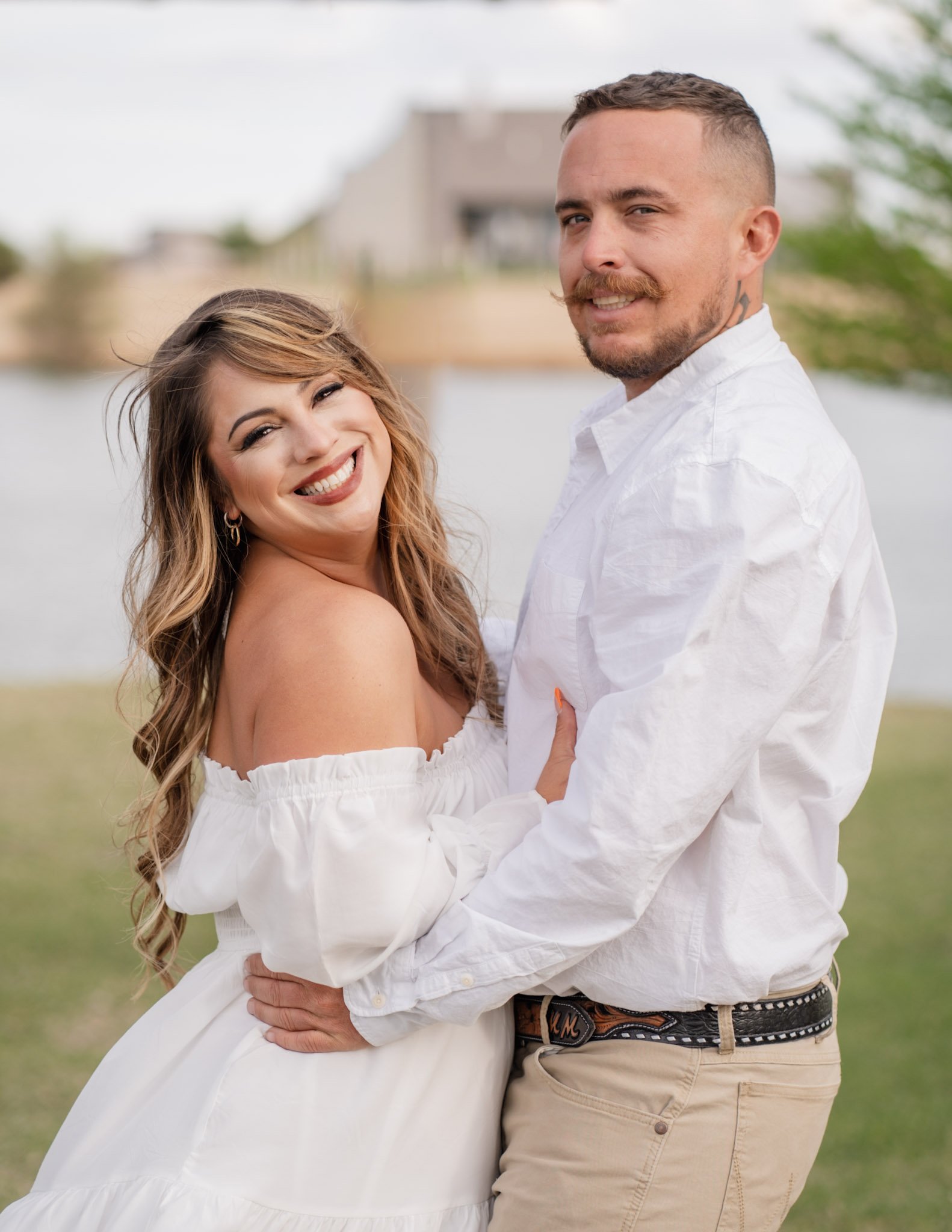 A smiling woman and a man holding each other outdoors on a grassy area with trees and buildings in the background.