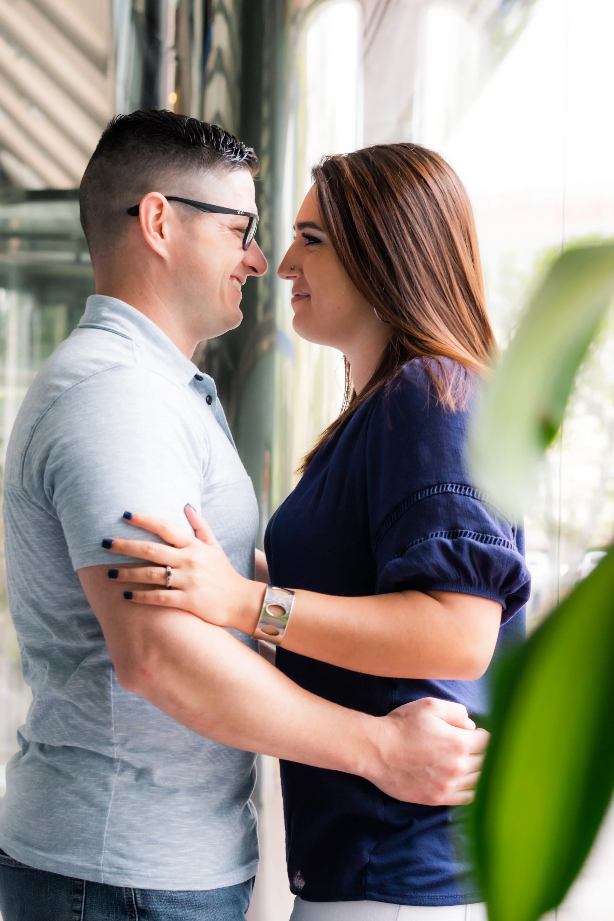 A young man and woman are smiling and looking into each other's eyes, holding each other close in a tender moment indoors near large windows.
