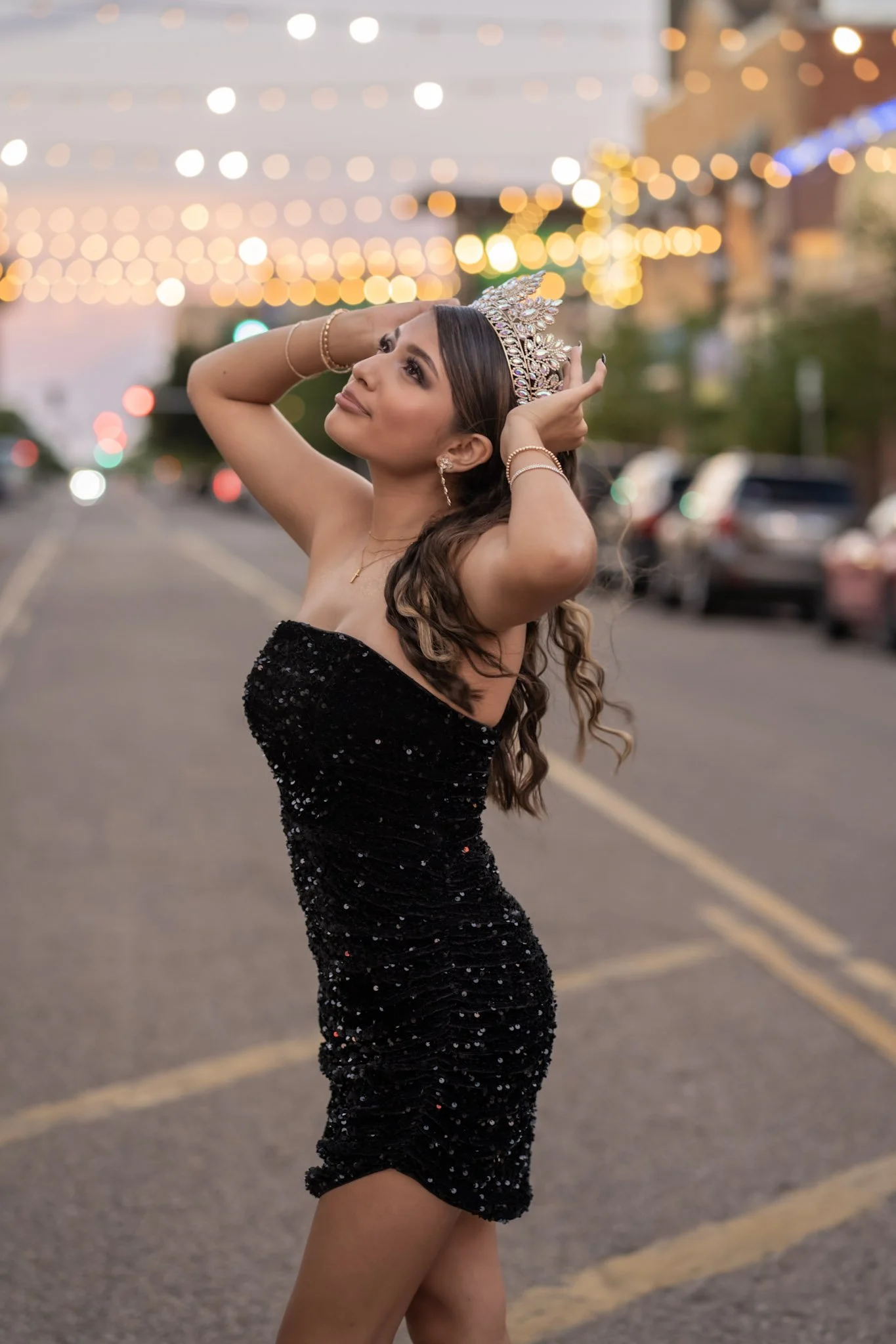 A woman in a strapless black sequined dress and wearing a glittering tiara poses with her hands on her head, standing on a city street at evening with blurry string lights in the background.