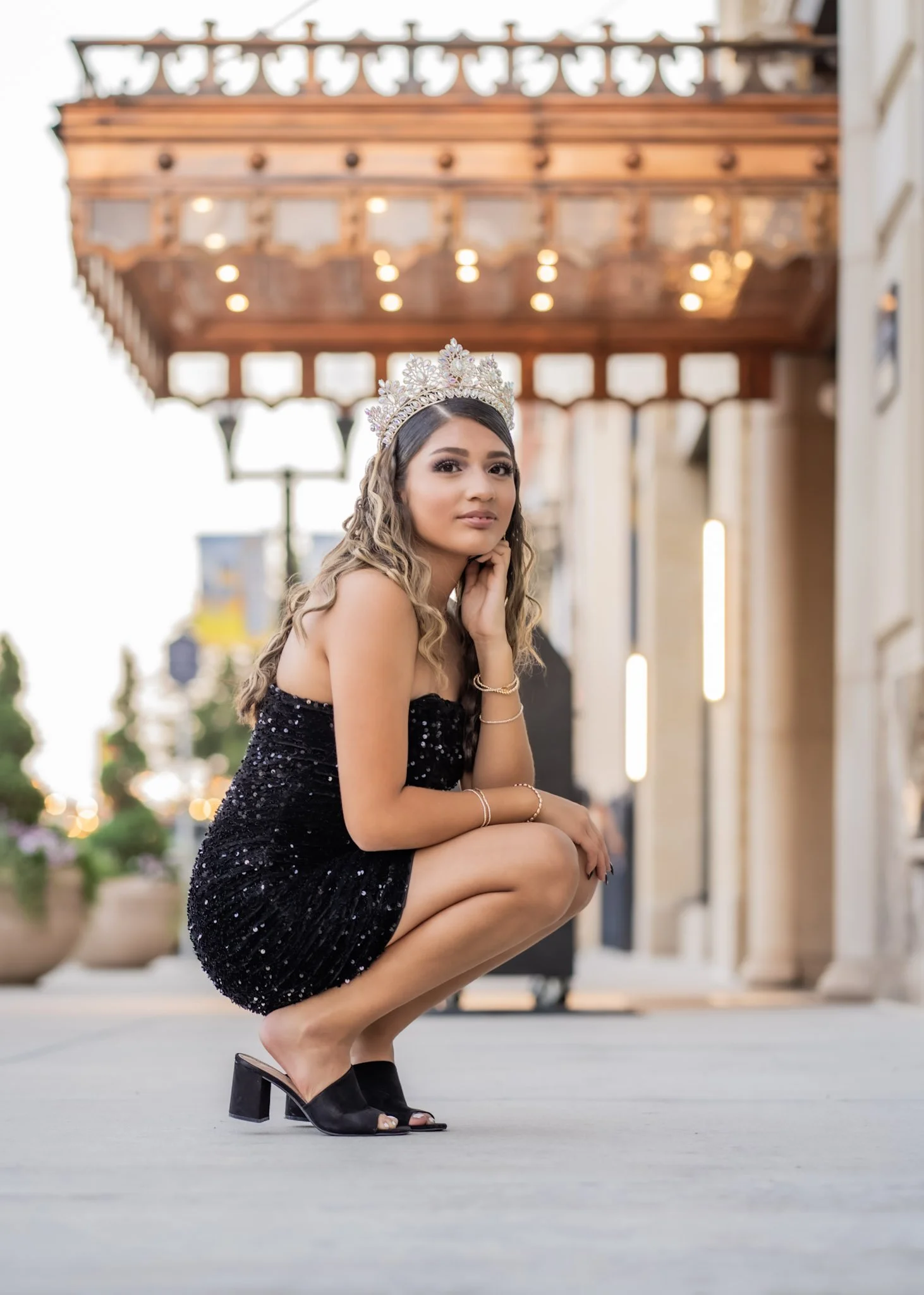 A young woman with long, wavy blonde hair wearing a black sequined dress, black heeled sandals, and a silver tiara, squatting outdoors in front of buildings with string lights, during evening or dusk.