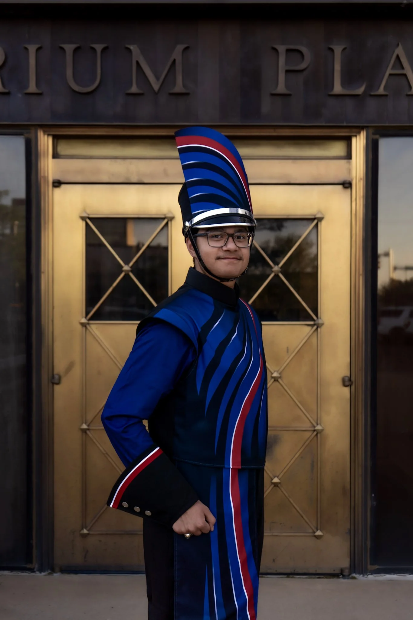 A young man in a band uniform standing in front of a gold-colored door with the word 'TRIUMPH' partially visible above. He is wearing a tall, colorful hat with red, white, blue, and black stripes, glasses, and has a confident expression.