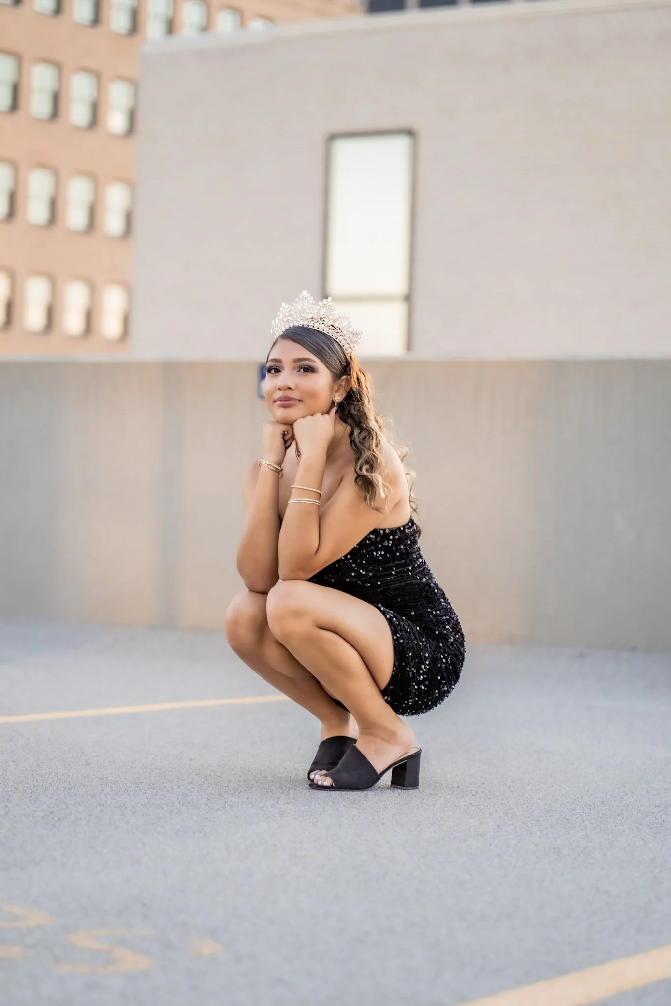 Young woman in a sparkly black dress with a tiara, squatting on an outdoor parking lot, with buildings in the background.
