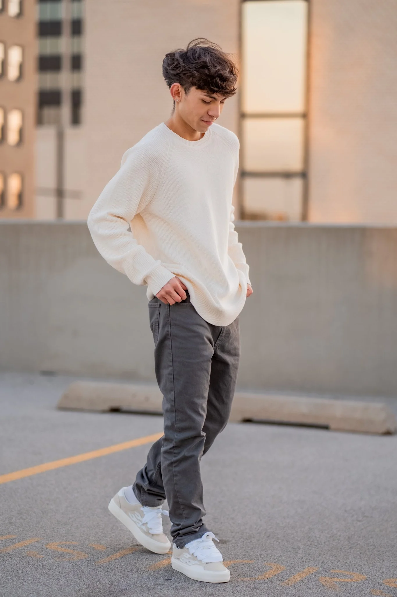 A young man with curly hair wearing a white sweater, dark gray pants, and white sneakers walking on a parking lot with painted lines.