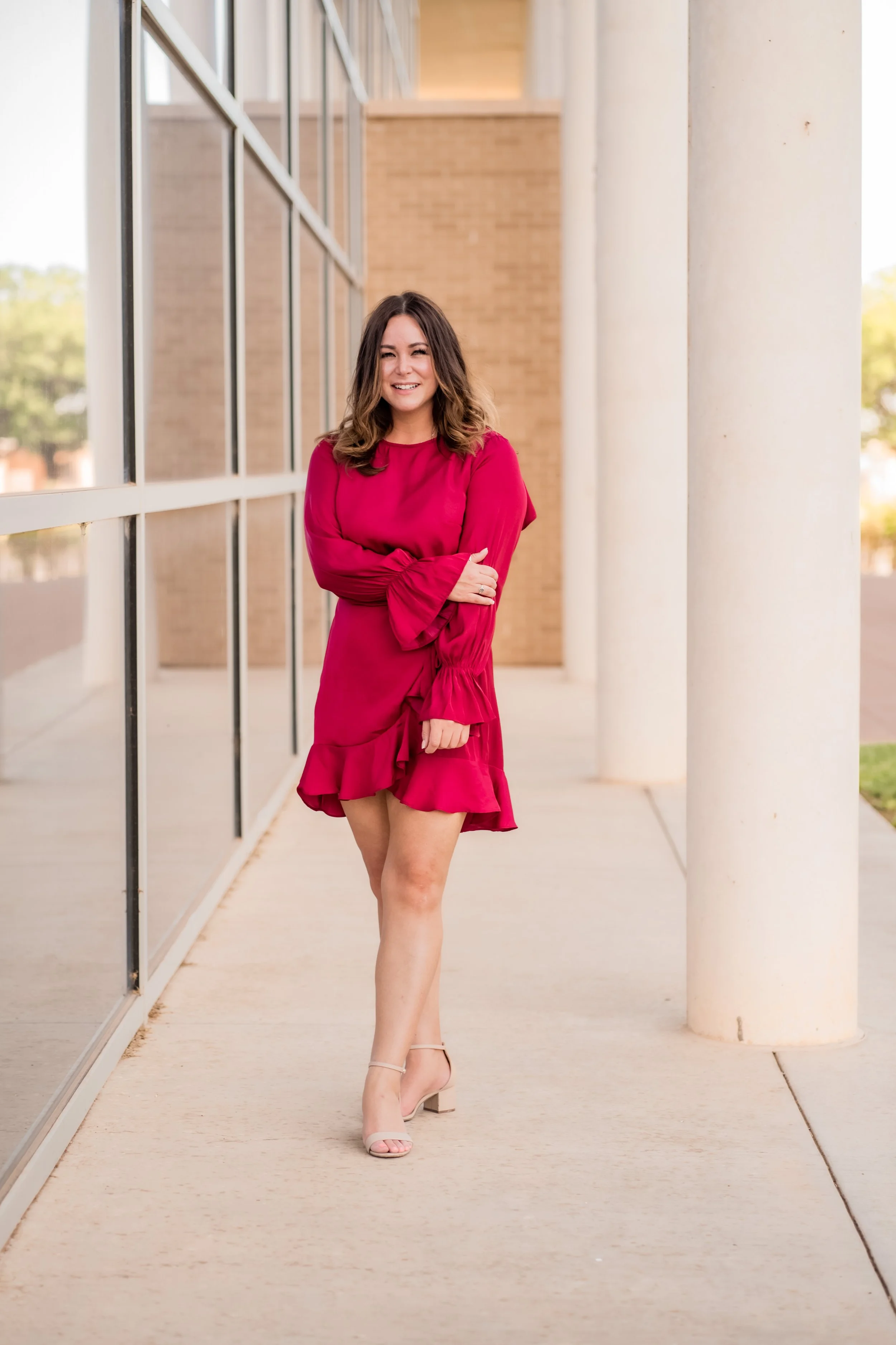 A woman with shoulder-length brown hair, wearing a bright pink dress with ruffled hem and long, flared sleeves, standing outside near a modern building with large glass windows and white columns, smiling and crossing her arms.