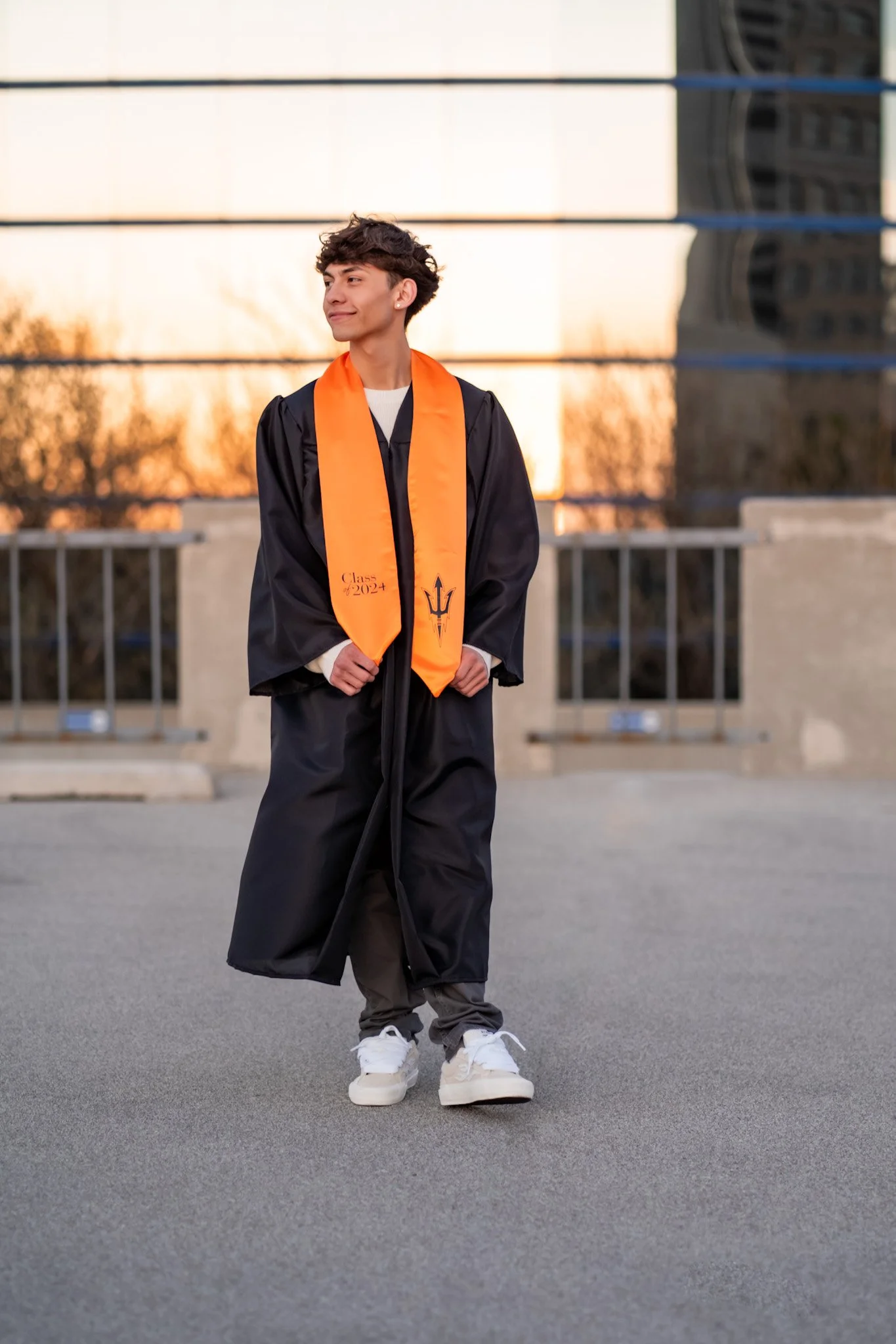 Young man in graduation gown and orange stole standing outdoors with a modern building reflecting on windows in the background during sunset.