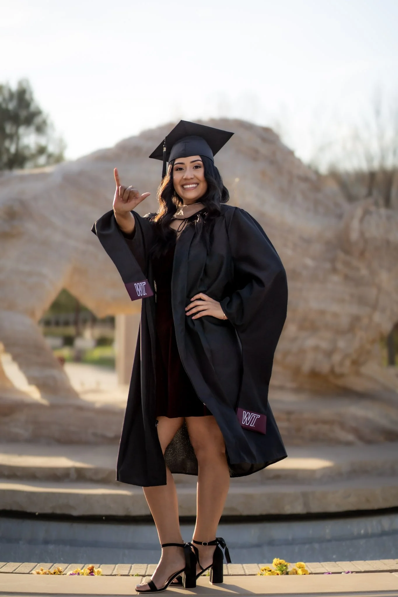 A young woman in a black graduation gown and cap is smiling and giving an L-shaped hand gesture outside during daytime. She is wearing high heels and is standing on a sidewalk in front of a brick building with flowers at her feet.