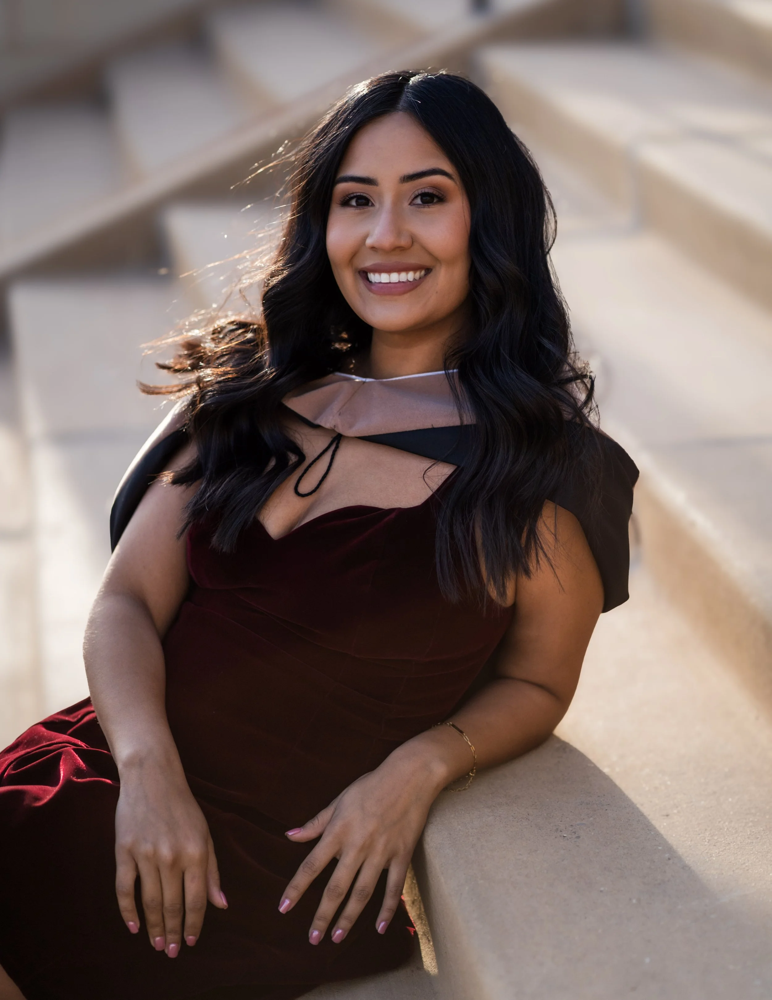 A woman with long dark hair smiling, wearing a burgundy dress with a sheer beige and black fabric around her shoulders, sitting on a sandstone structure outdoors during golden hour.