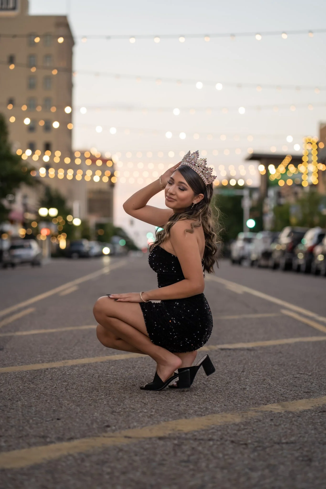 A woman wearing a black dress with sparkles and high heels kneels on a city street at dusk, wearing a crown and smiling gently with her eyes closed. String lights and buildings are visible in the blurred background.