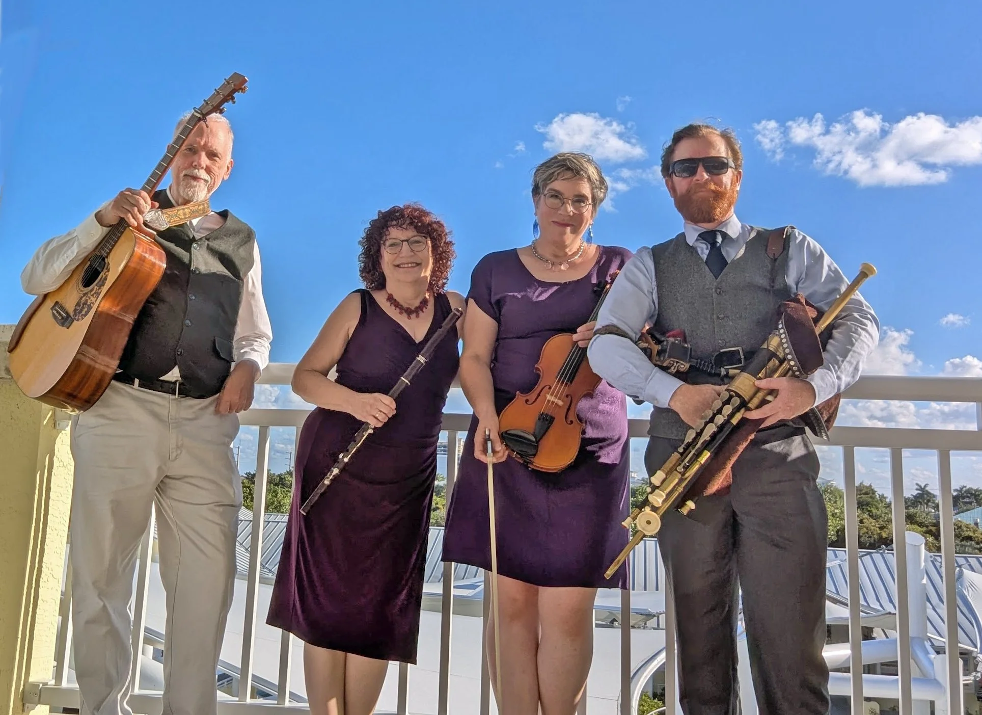 Four Irish musicians standing in front of a blue sky in Florida. The musicians from left to right are: Andy Thurston, guitar, Elise Kress, Irish flute, Julia Fitzgerald, Fiddle and Stewart Pittman, Uilleann pipes.