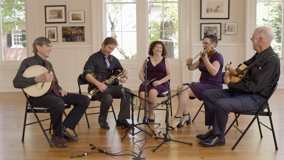 Five musicians are seated in a semicircle in a beautiful building in Alexandria, Virginia. From left to right are: Jesse Winch, Bodhran, Stewart Pittman, Uilleann pipes, Elise Kress, penny whistle, Julia Fitzgerald, fiddle and Andy Thurston, guitar.