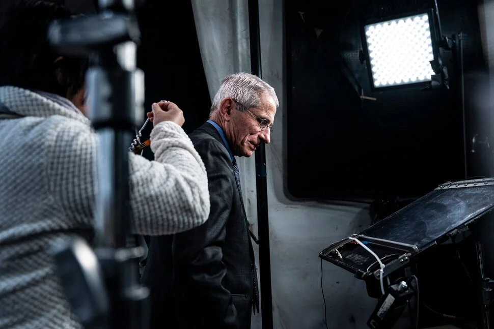 A man with gray hair and glasses, dressed in a suit, appears to be on a film or video set, leaning forward and looking at a monitor.