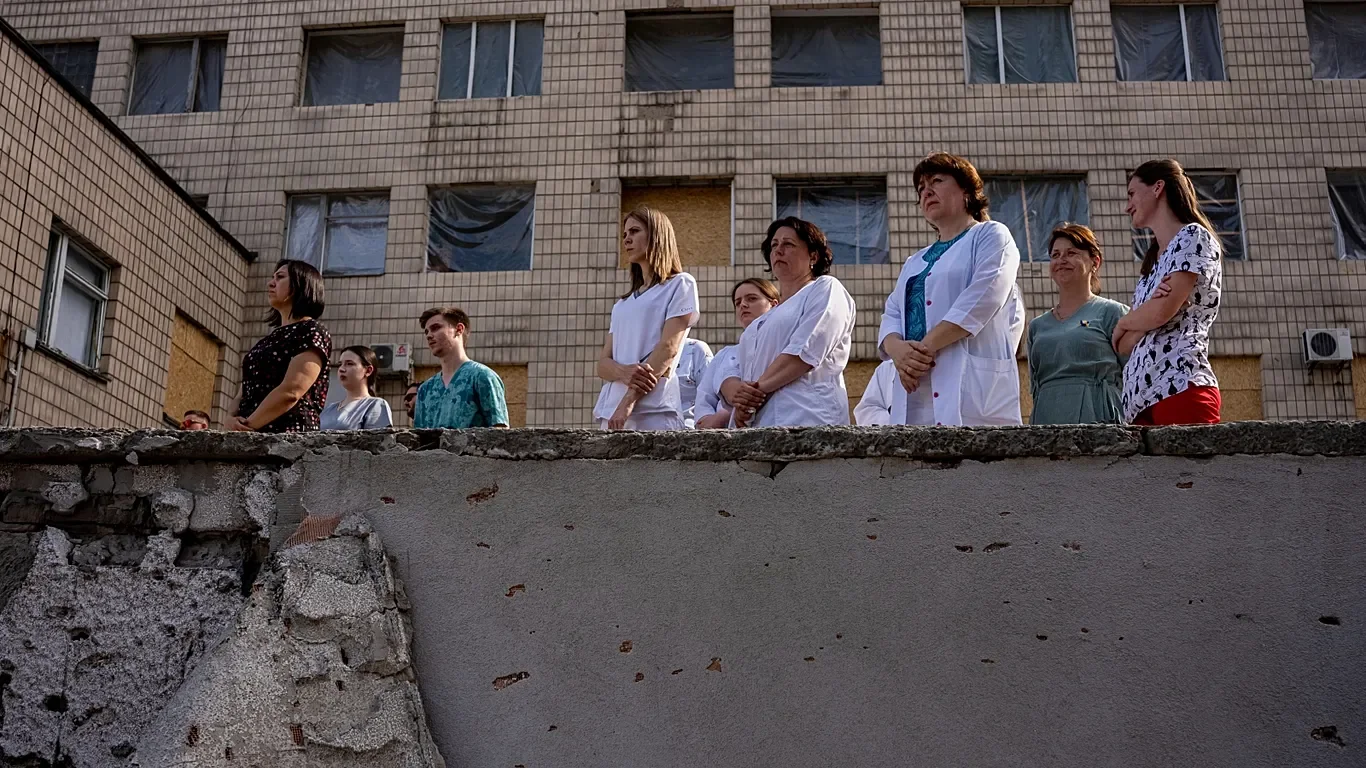 Group of medical professionals, including nurses and doctors, standing outside a building with boarded-up windows, observing something below.