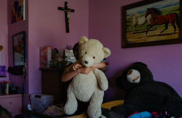 A girl holding a large teddy bear in a purple-walled room decorated with horse paintings and a cross on the wall.