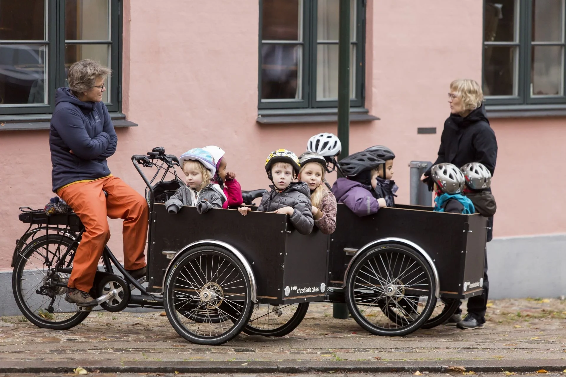 A man riding a bike with a cargo bike attached, carrying six young children all wearing helmets, two women standing nearby, in front of a pink building with multiple windows.