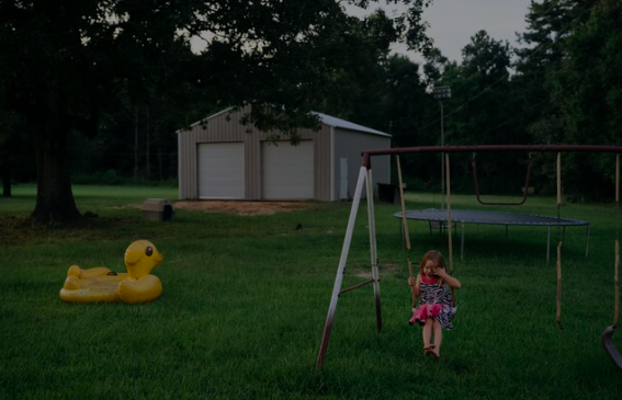 A young girl sitting on a swing set in a backyard with a trampoline, a yellow rubber duck float, and a shed in the background.