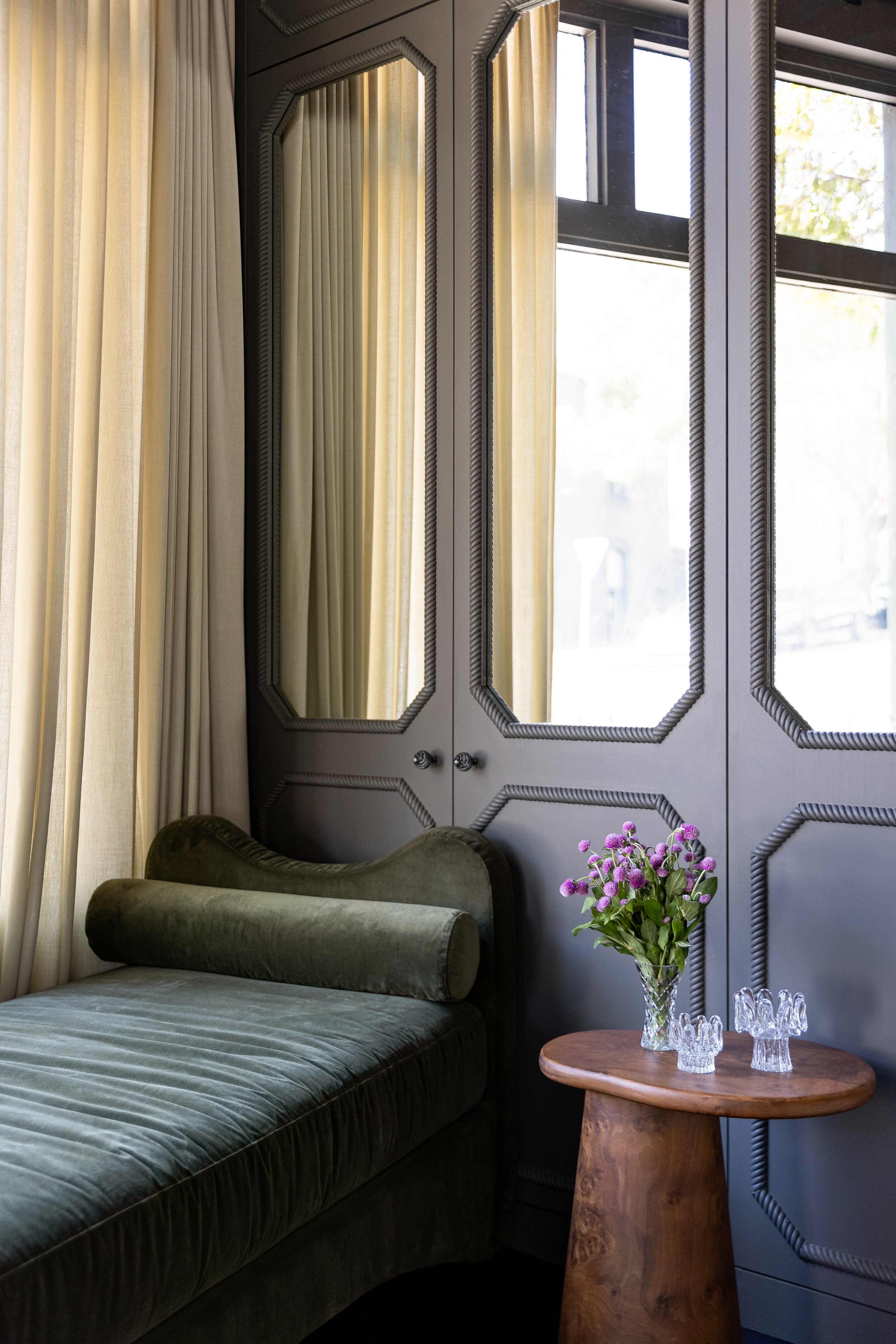 A cozy corner with a green velvet chaise lounge, a wooden side table with a vase of purple flowers, and a wall with decorative black paneling next to large windows with cream curtains. SMAC Studio Photo by Ruth Gold