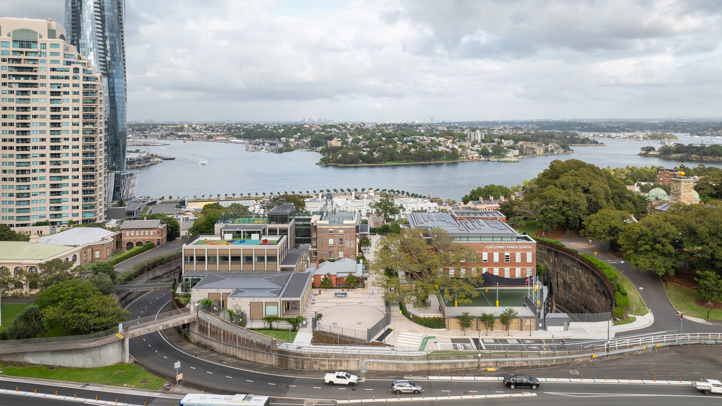 A cityscape view featuring high-rise buildings, a river or bay with boats, trees, and a school called Fort Street Public School with sports facilities in an urban area. Designed by fjcStudio Photo by Ruth Gold