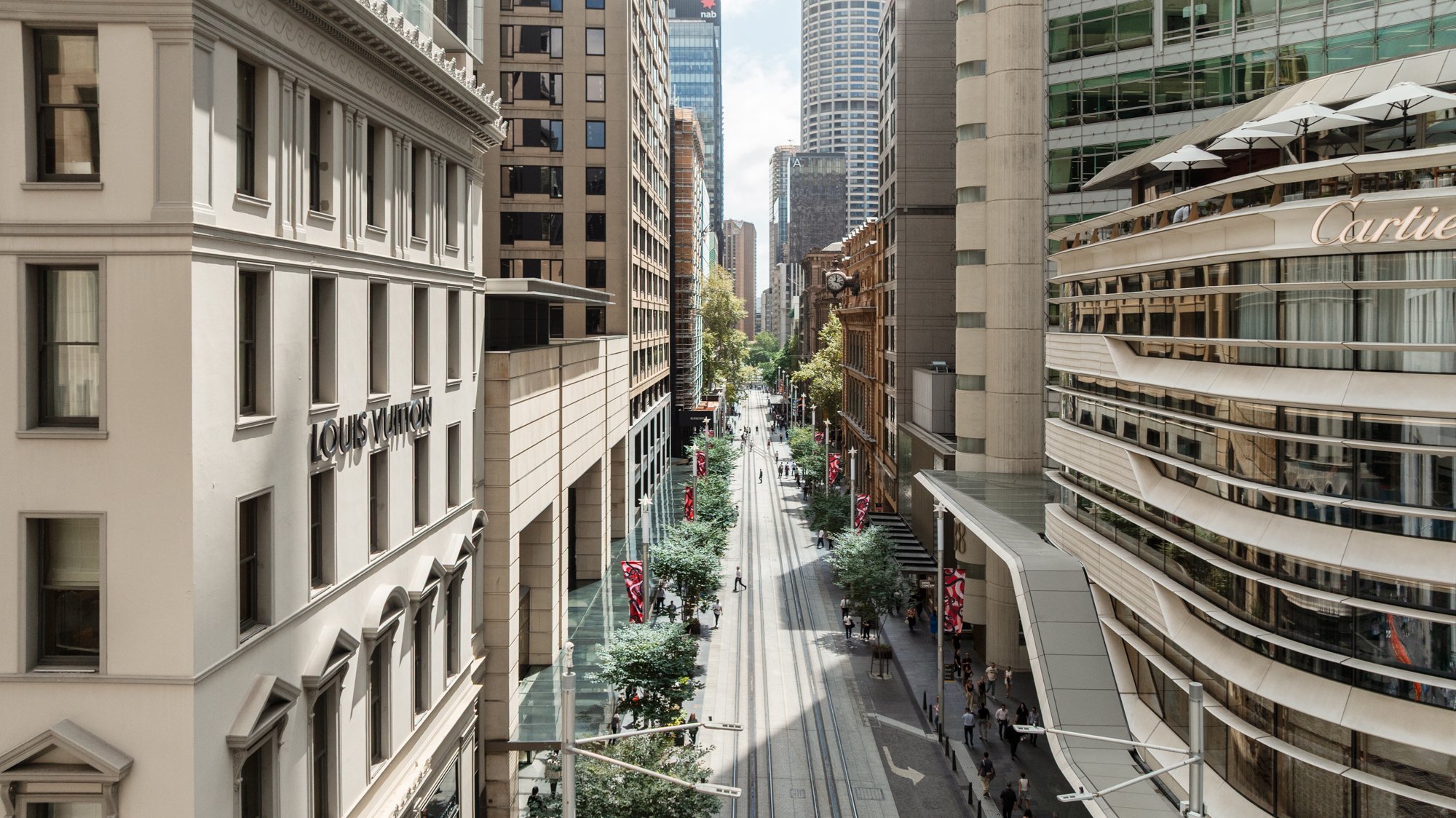 A city street viewed from above with tall buildings on both sides, storefronts, trees lining the sidewalks, and a few pedestrians walking. Designed by fjcStudio Photo by Ruth Gold