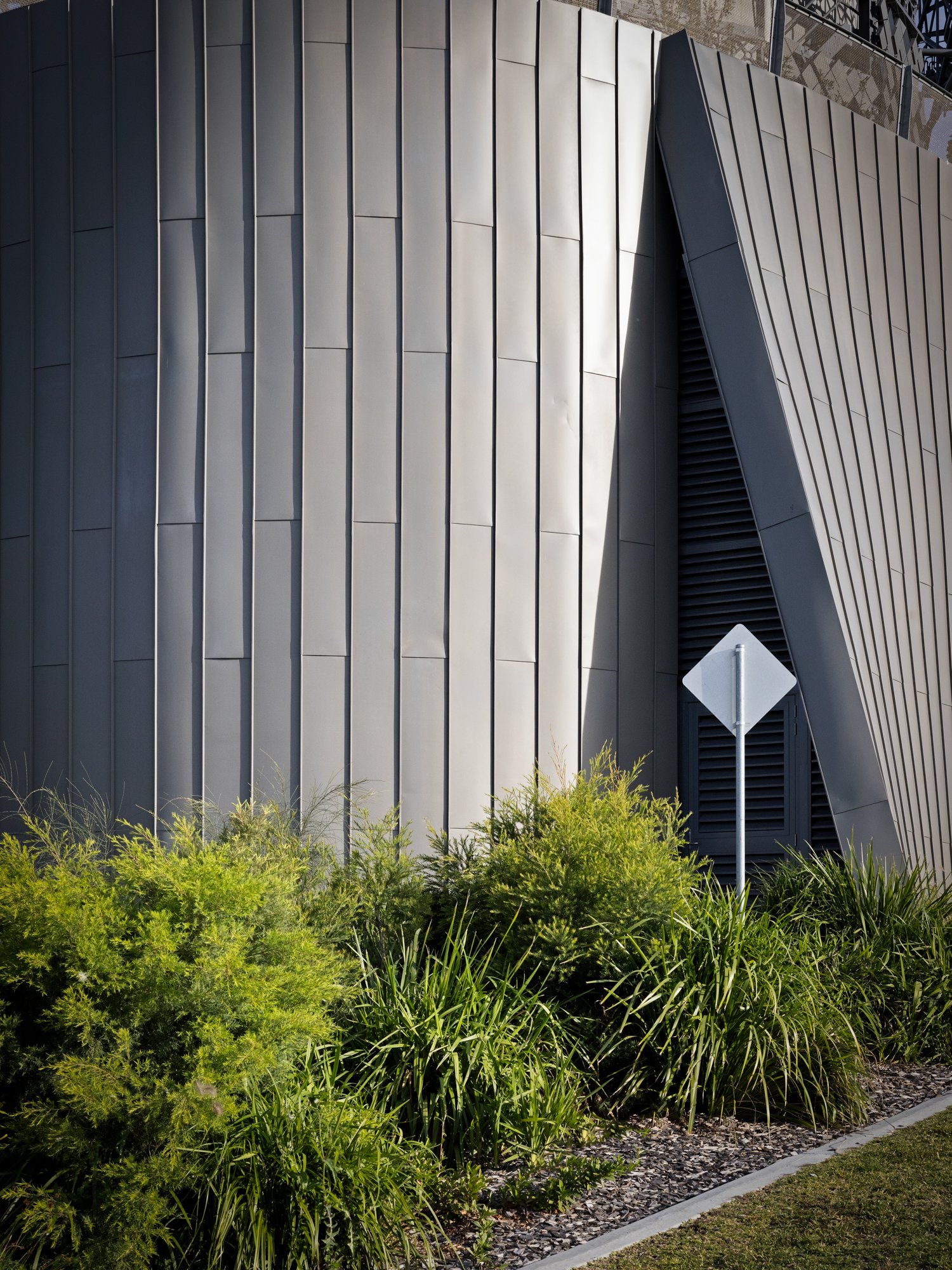 Modern building with metallic panels, black vent, and a white street signpost in front of greenery. Designed by Colin Polwarth Studio Photo by Ruth Gold