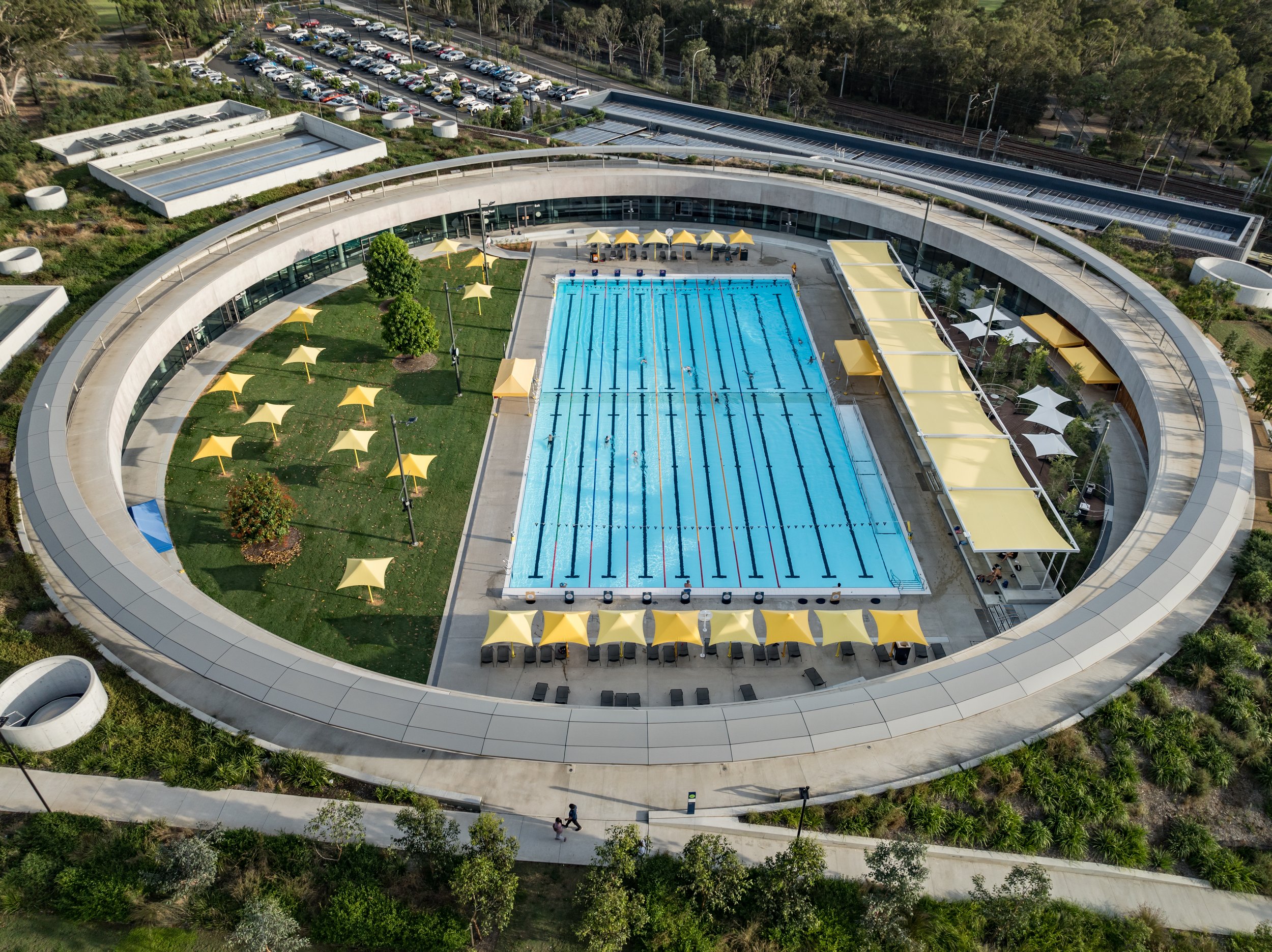 Aerial view of an outdoor swimming pool area surrounded by a modern, circular building with yoga or lounge areas, yellow umbrellas, and surrounding greenery. Designed by McGregor Coxall Photo by Ruth Gold