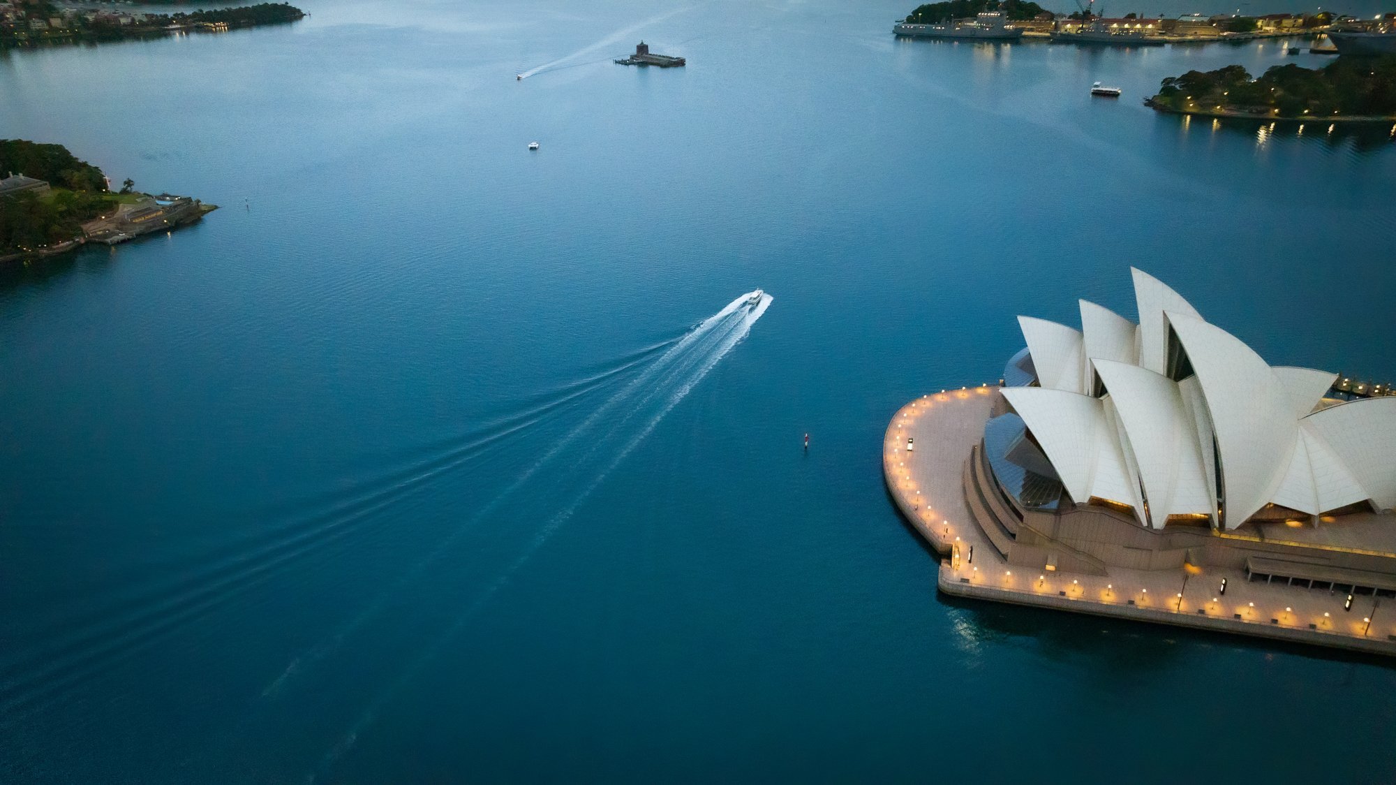 Aerial view of Sydney Opera House with a boat leaving a wake in the water, nearby land with buildings and greenery, and calm water reflecting the sky. Opera House Photo by Ruth Gold