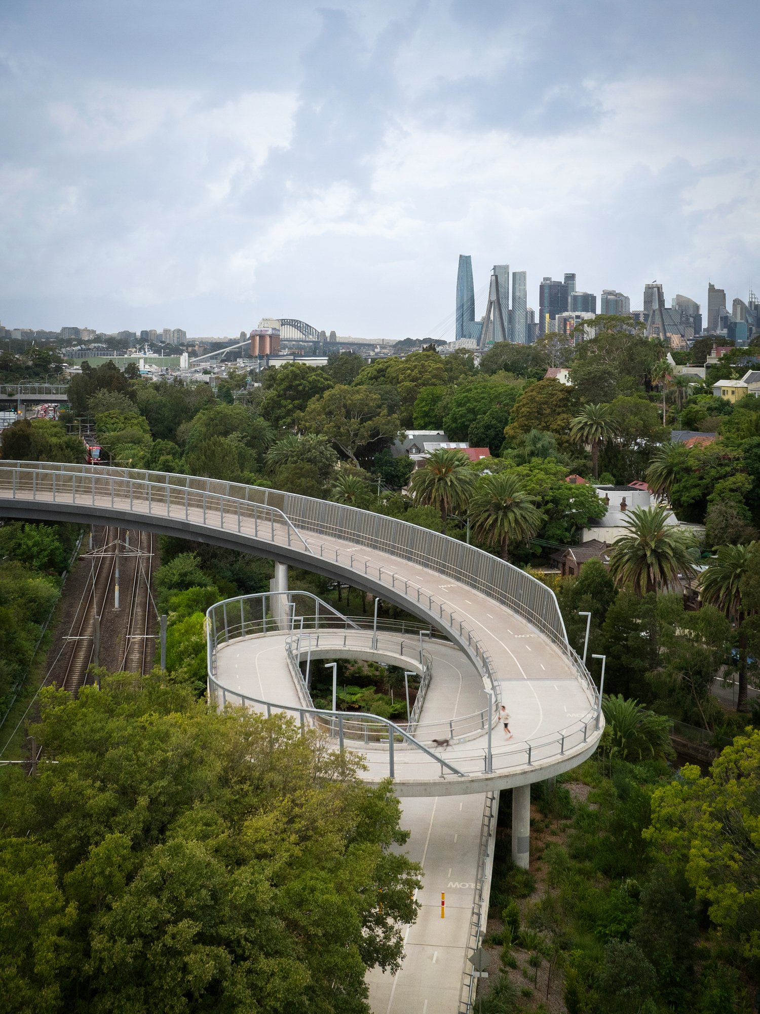 A winding elevated walkway in a park with lush green trees, set against a city skyline with tall buildings and cloudy sky in the background. Designed by Colin Polwarth Studio Photo by Ruth Gold