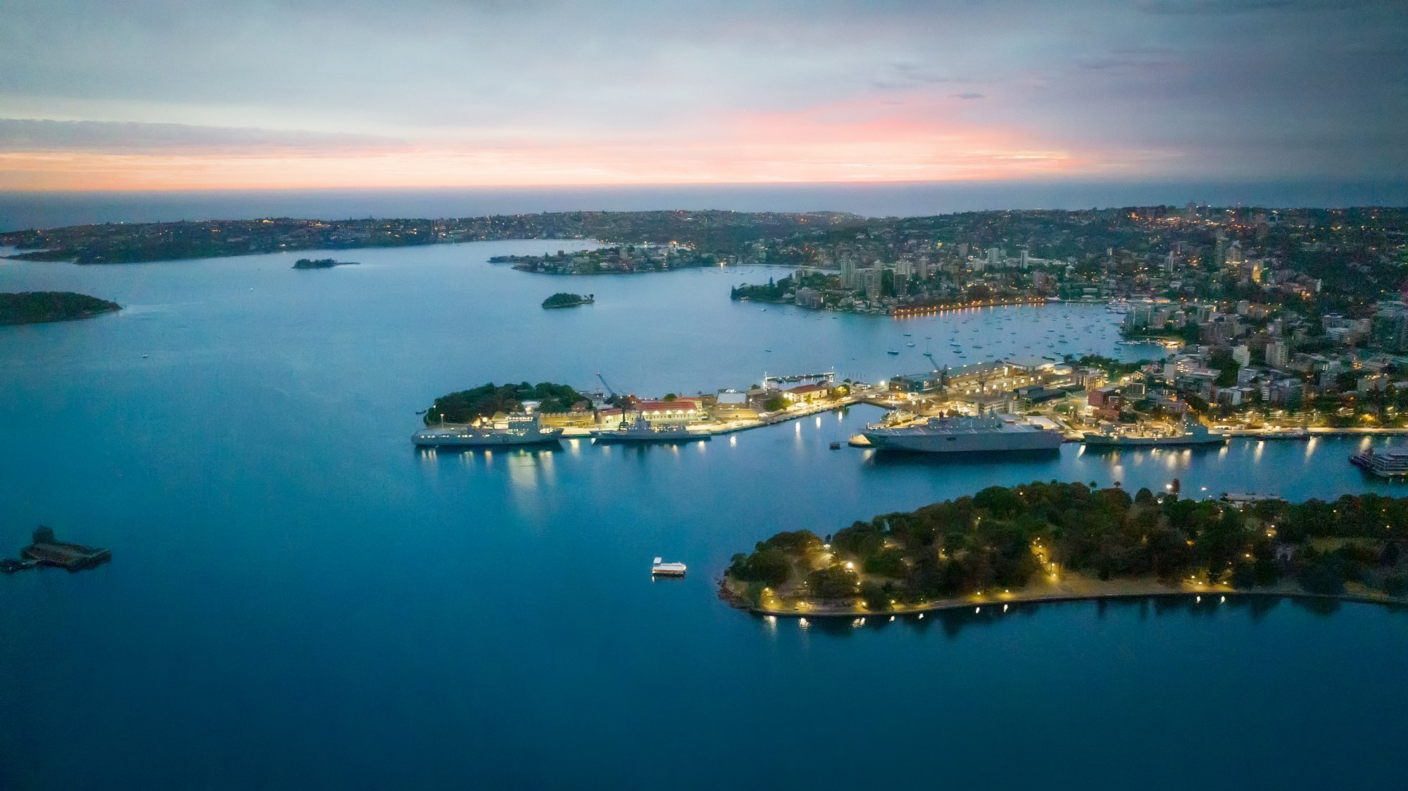 An aerial view of a harbor at dusk with boats docked and city lights illuminating the shoreline. Opera House Photo by Ruth Gold