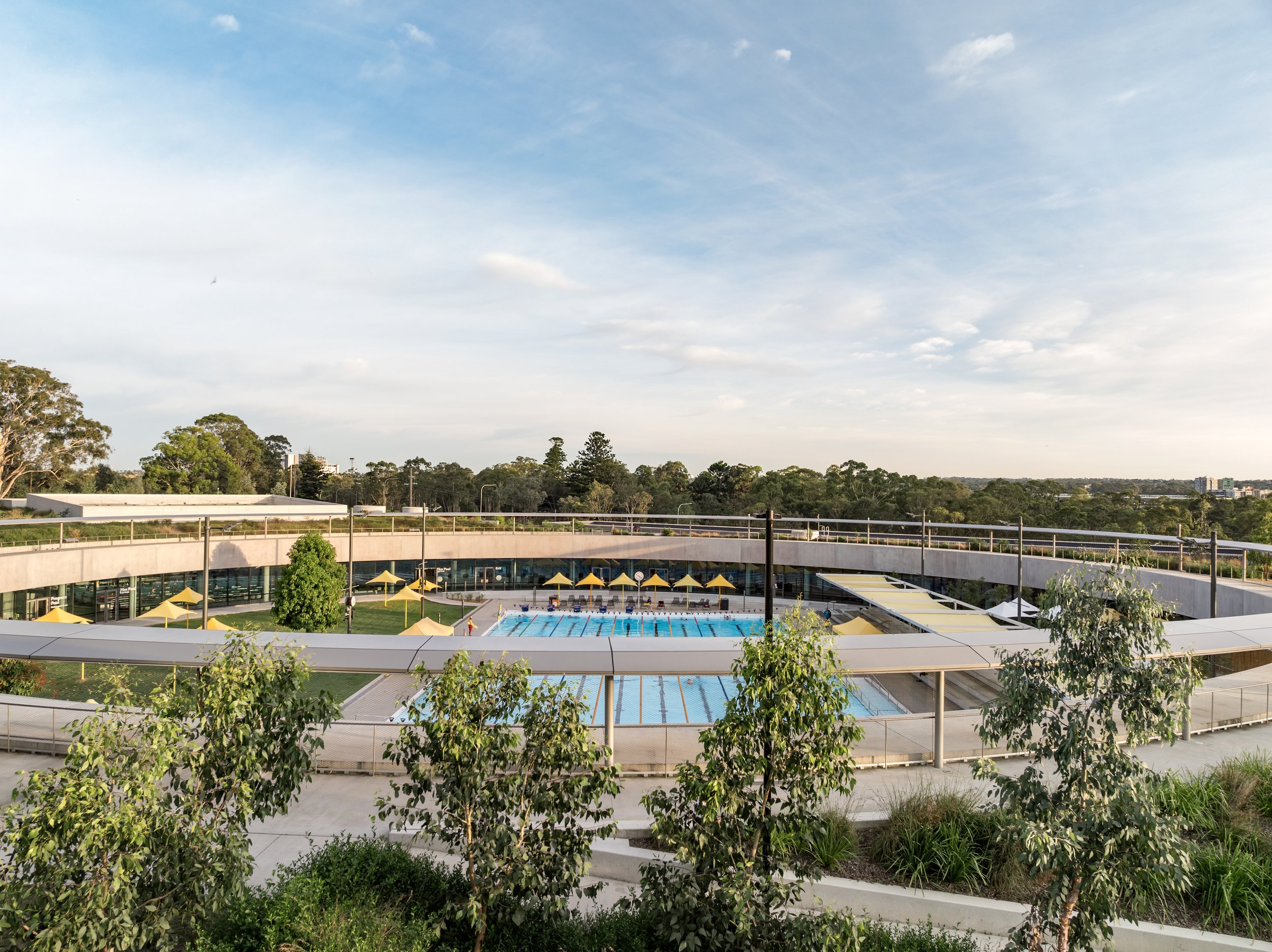 Outdoor swimming pool area with lane swimming, surrounded by yellow umbrellas, trees, and greenery, under a partly cloudy sky. Designed by McGregor Coxall Photo by Ruth Gold